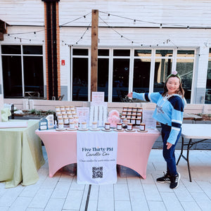 Ivy, founder and candle maker of Five Thirty PM Candle Co®, standing next to a market display featuring Glow Up and Archive of Life candles and wax melts at Ballston Pop-up Market.