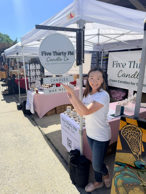 Ivy of Five Thirty PM Candle Co® standing beside her branded outdoor makers market booth displaying handcrafted candles and wax melts.