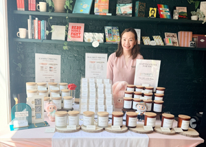 Ivy, founder and candle maker of Five Thirty PM Candle Co®, standing behind a market display featuring Glow Up and Archive of Life candles and wax melts.