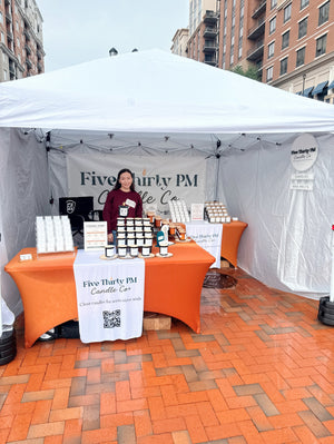 Ivy, founder and candle maker of Five Thirty PM Candle Co®, standing inside her outdoor market booth with displays of handcrafted candles, wax melts, and candle accessories at Anchors & Artisans September market hosted by the Makers of Maryland
