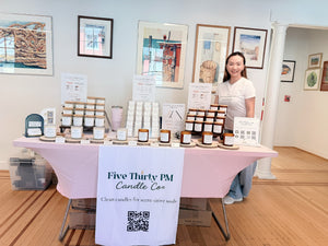 Ivy, founder and candle maker of Five Thirty PM Candle Co®, standing behind a market display featuring Glow Up and Archive of Life candles and wax melts at The Market at Caplan's.